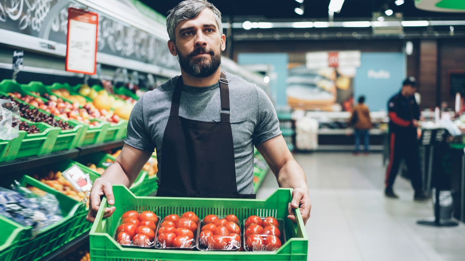 A grocery store worker in an apron carries a crate of tomatoes through the produce section.
