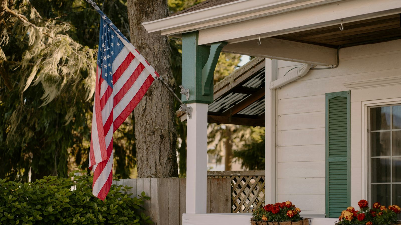 An American flag hangs from the porch of a white house with green shutters and flower boxes.