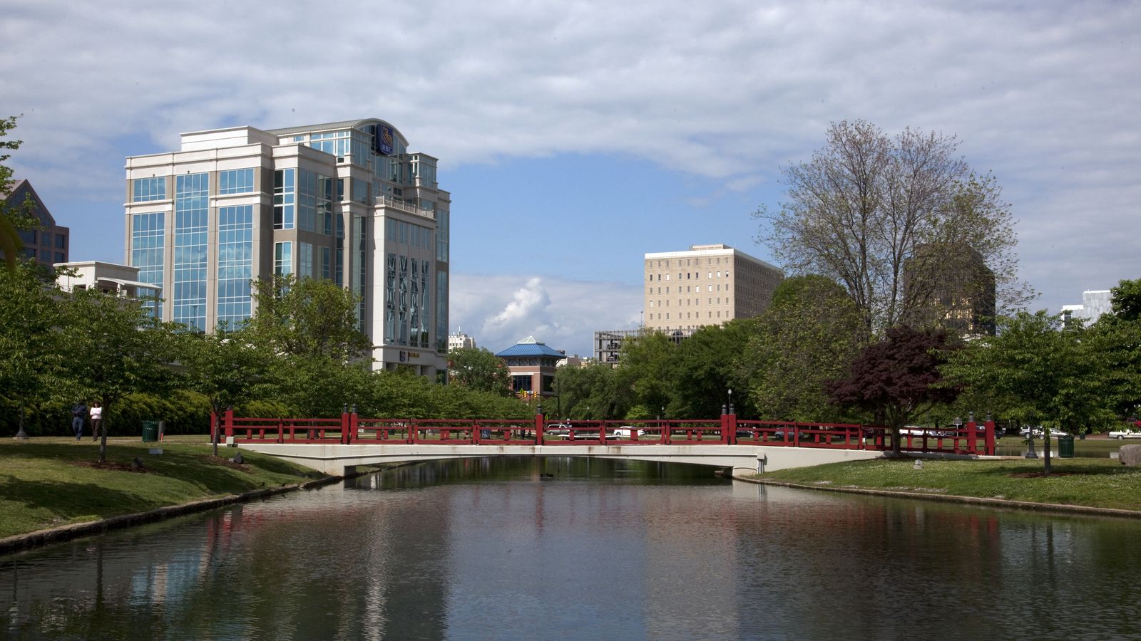 Big Spring Park in downtown Huntsville, with a red footbridge over the pond and a glass office tower behind the trees.